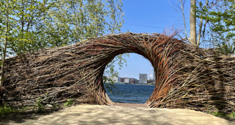 Een rond gewoven takkenportaal met zicht op water en stad in de verte.