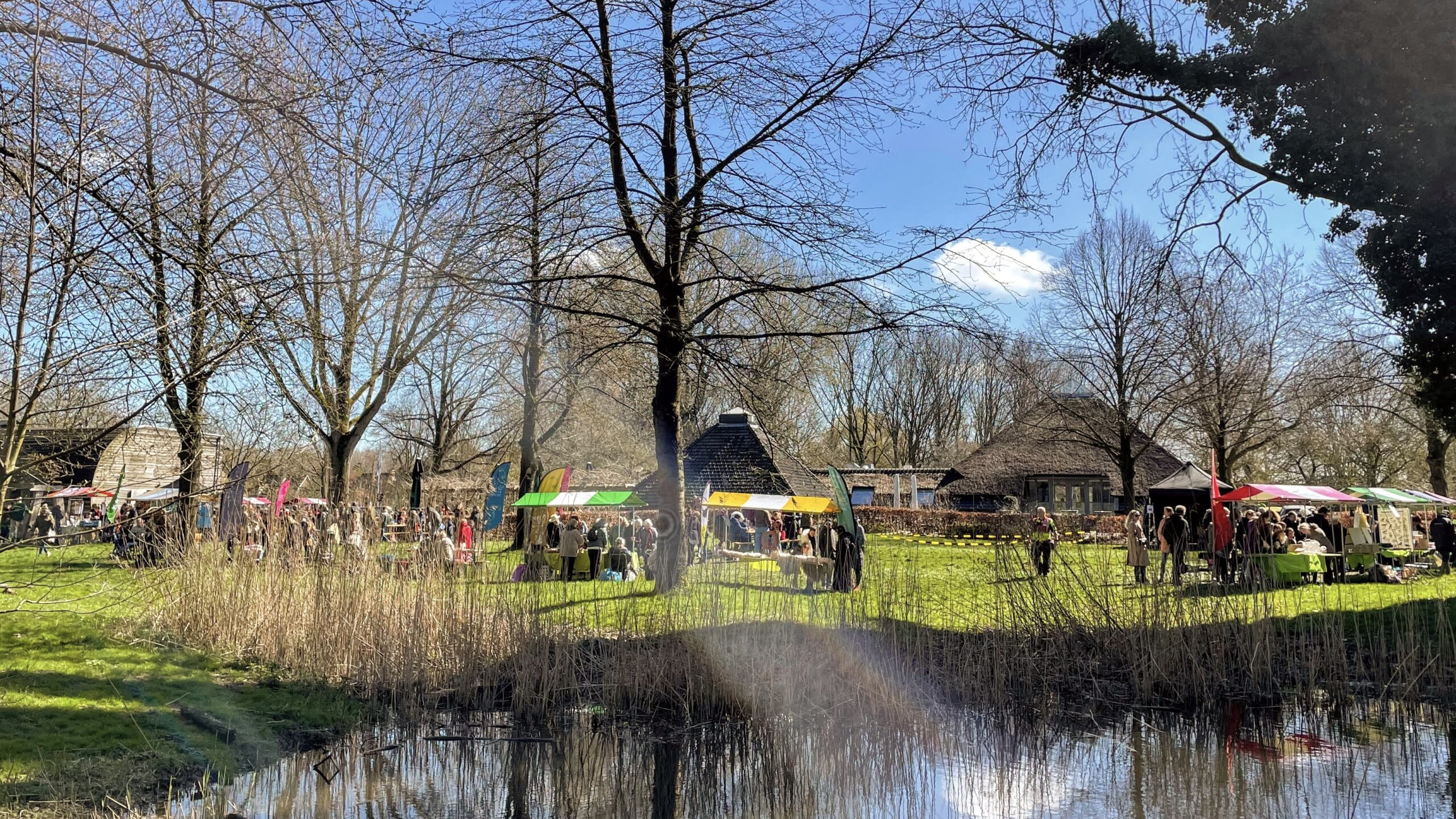 Buitenmarkt met kraampjes in een park; mensen wandelen onder kale bomen bij een vijver.
