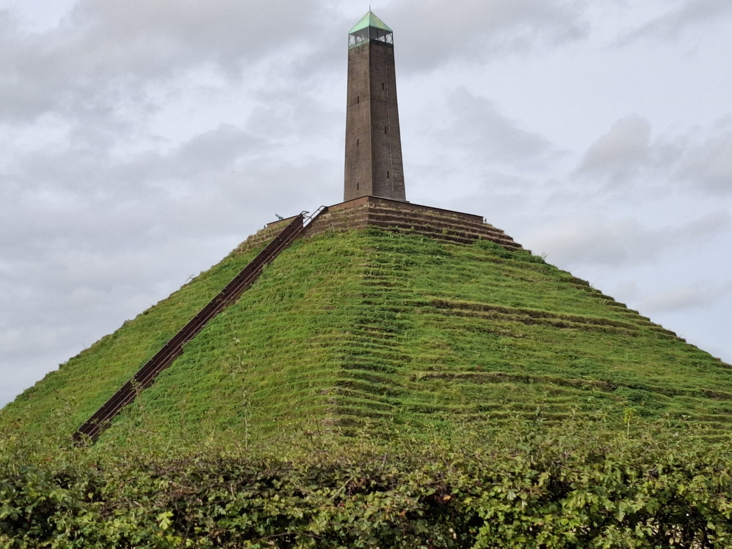 Groene piramidevormige heuvel met een stenen toren bovenop, plus een rechte trap aan de zijkant.