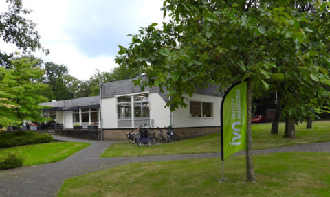 Wit gebouw met IVN-vlag en fietsenrekken op grasveld met bomen.
