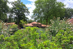Groene tuin met bloeiende planten voor een gebouw met rode dakpannen, omgeven door bomen.