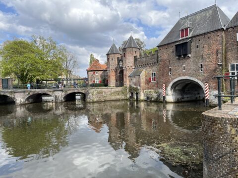 Middeleeuwse poort en brug over water, met reflecties en groene bomen, onder een bewolkte hemel.