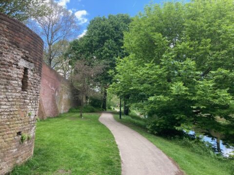 Een wandelpad langs een oude bakstenen muur en groene bomen op een zonnige dag.