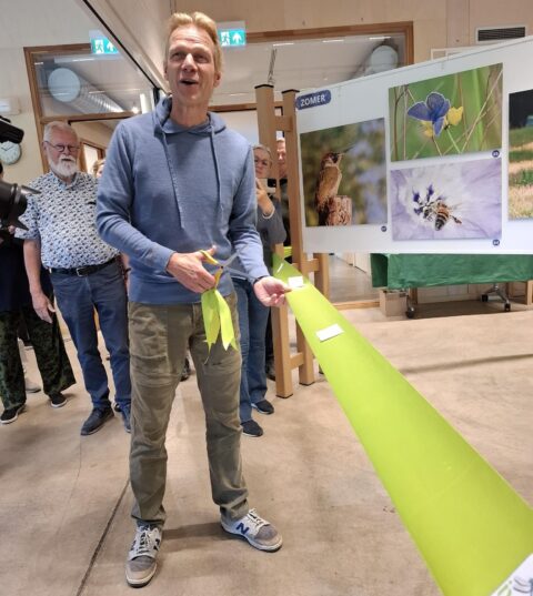 Man knipt groen lint door, omringd door mensen. Fotopanelen met natuurfoto’s op de achtergrond.