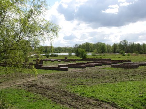 Ruïnes met stenen funderingen in een groen landschap naast een rivier onder een bewolkte hemel.