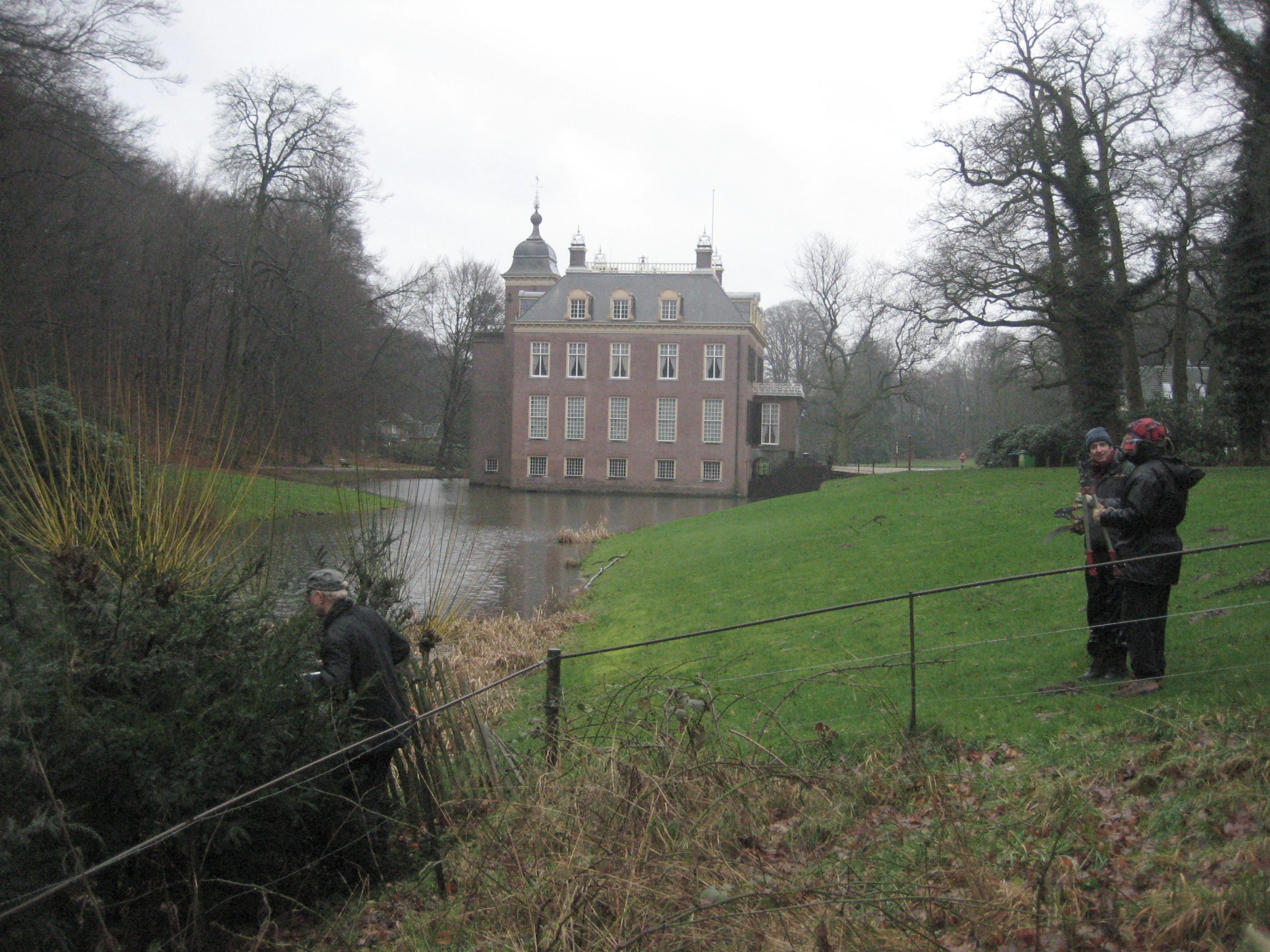 Kasteel met gracht, winterse bomen en mensen in regenjassen op grasveld.