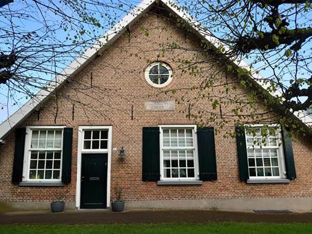 Bakstenen huis met drie ramen, groene luiken en een rond raam onder de dakrand. Grasveld en bomen.