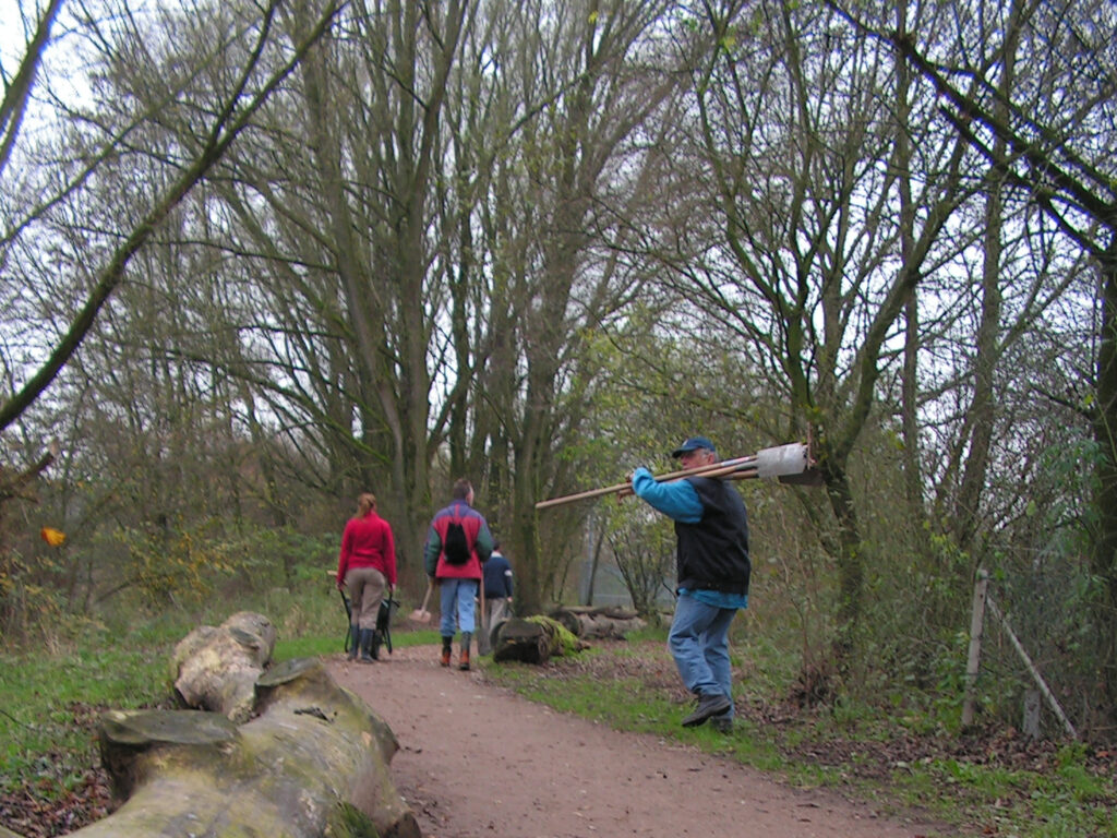 Mensen wandelen in een bosrijke omgeving met een persoon die houten stokken draagt.