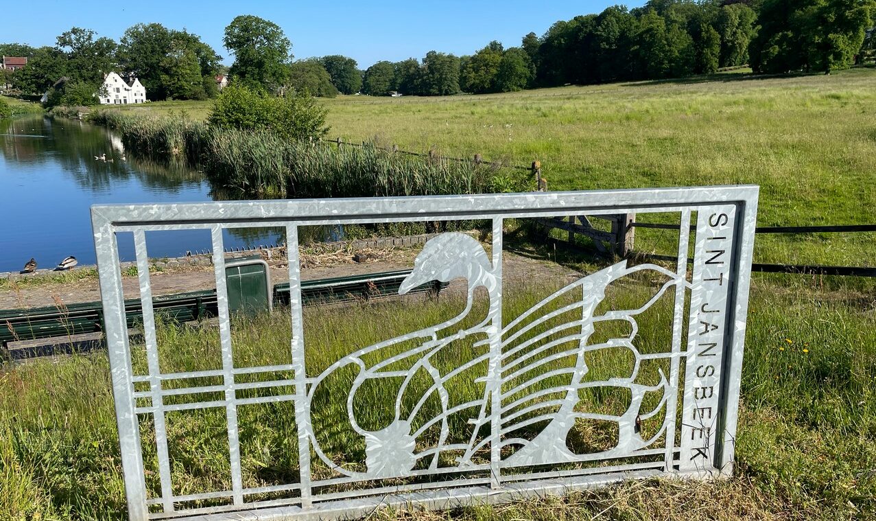 Metalen hek met zwanenpatroon en tekst "Sint Jansbeek" voor een landschap met gras en water.