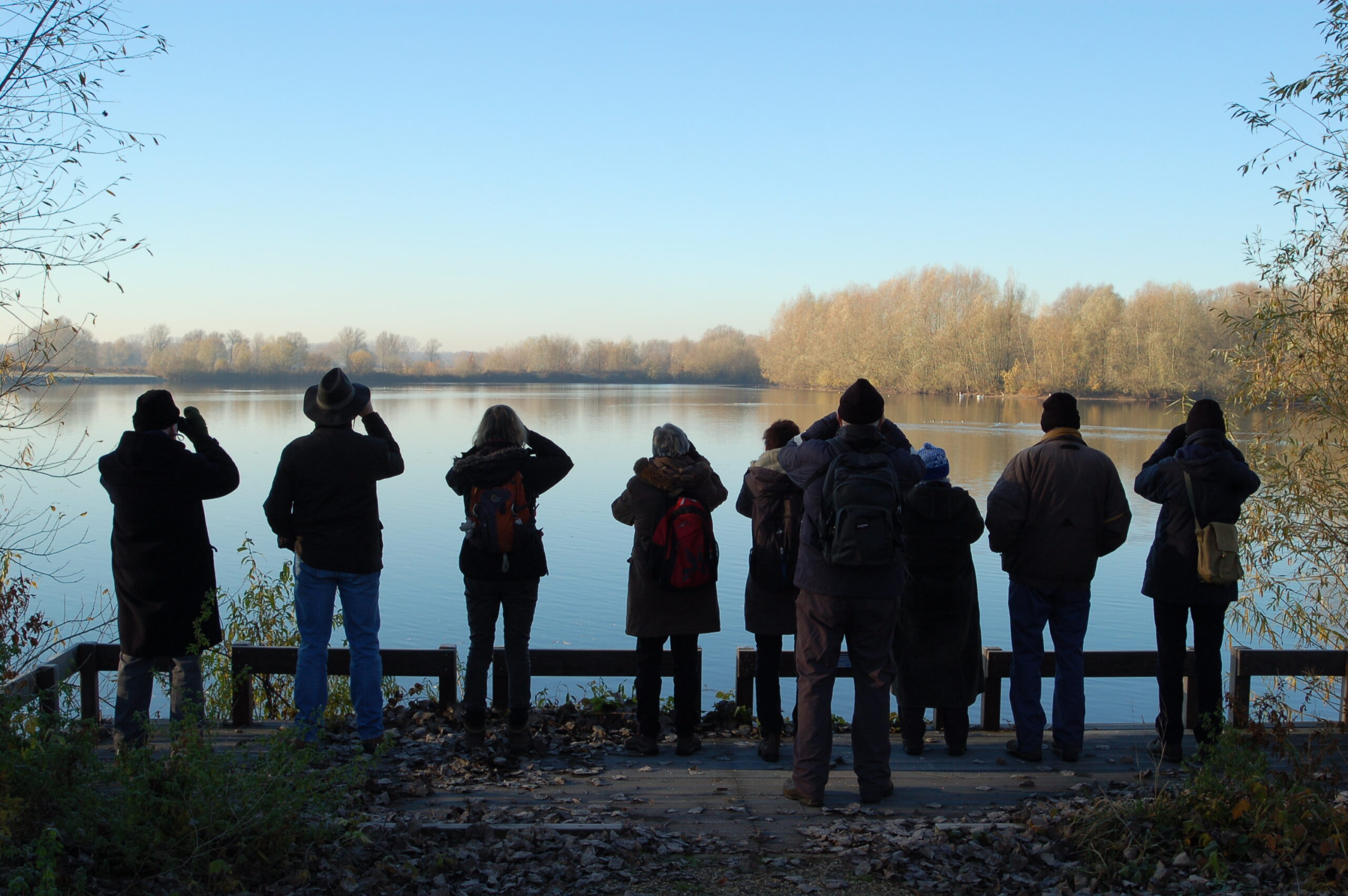 Groep mensen kijkt met verrekijkers naar een meer met bomen op de achtergrond.