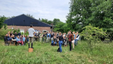 Een man spreekt buiten tot een groepsbijeenkomst bij een landelijke boerderij.