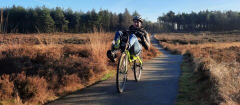 Persoon fietst op een ligfiets op een smal pad door een bosrijke, open heide met een blauwe lucht.