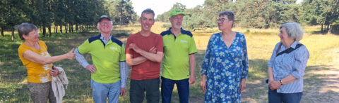 Groep van zes mensen staat buiten in een bosrijke omgeving, sommigen dragen groene shirts.