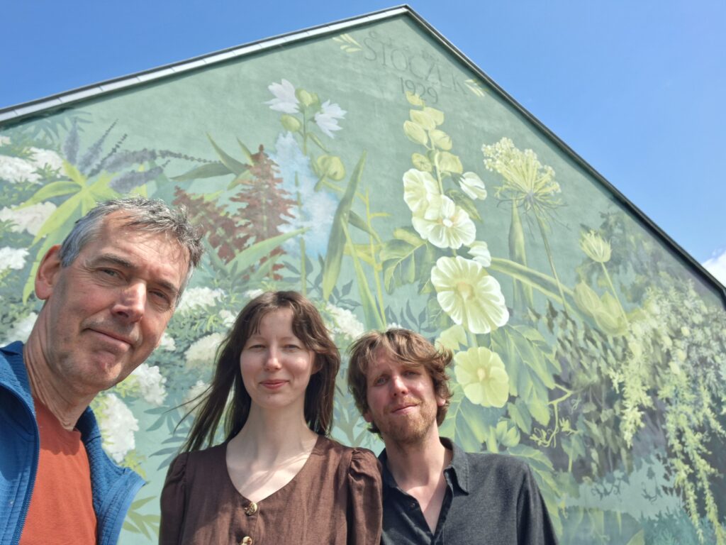 Drie mensen voor een muurschildering met bloemen en planten, onder blauwe lucht.
