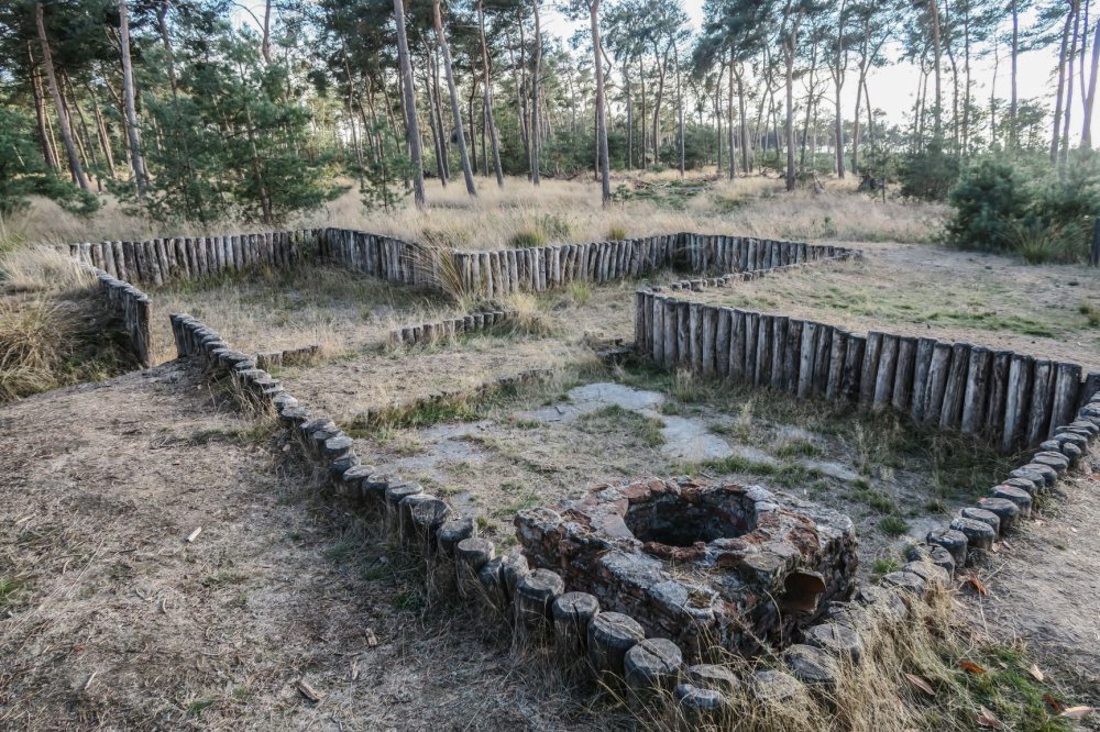 Houten omheining vormt een vierkant patroon met een stenen put, omgeven door boslandschap.