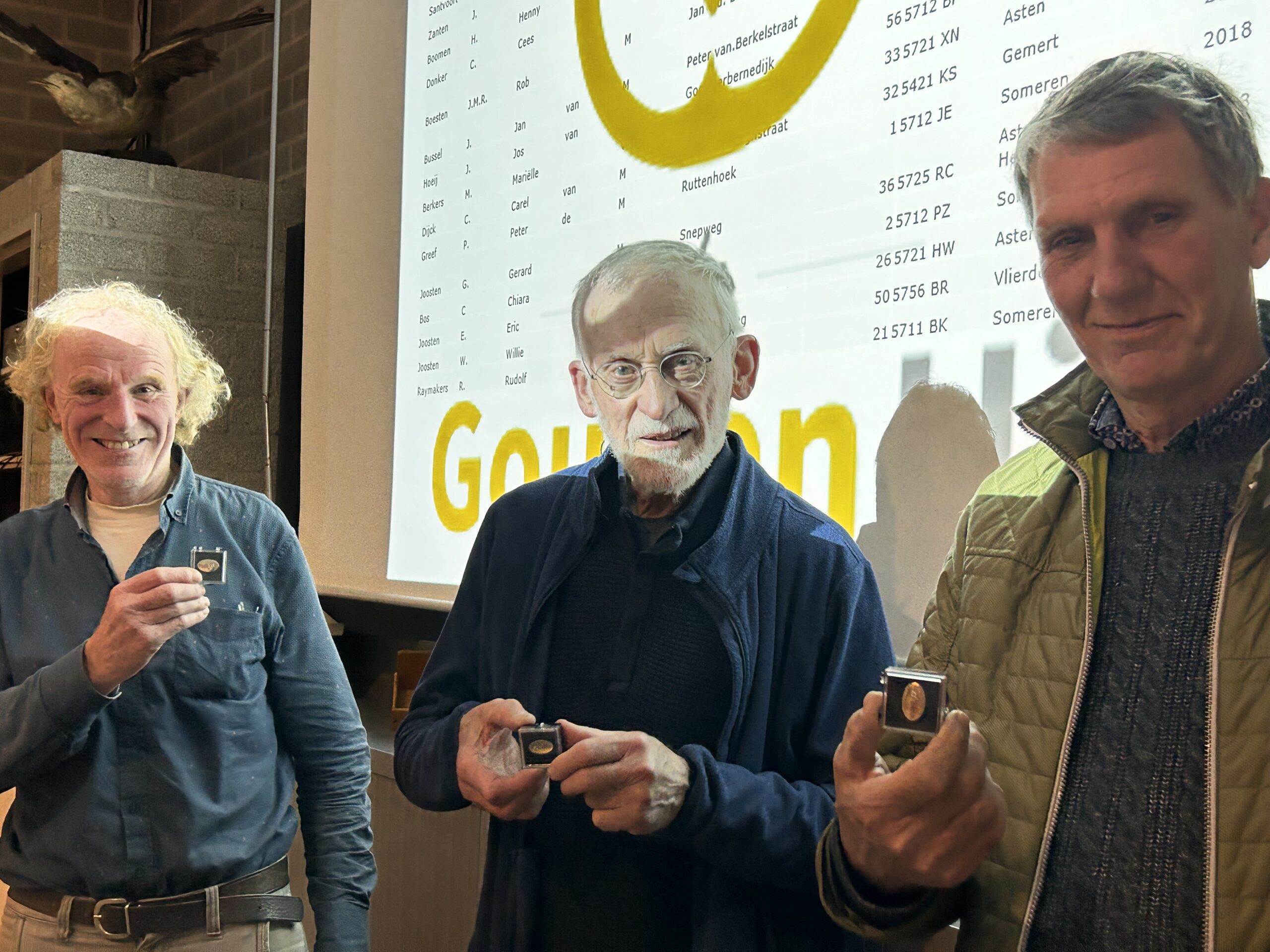 Drie mannen poseren met medailles voor een scherm met namen en tekst.