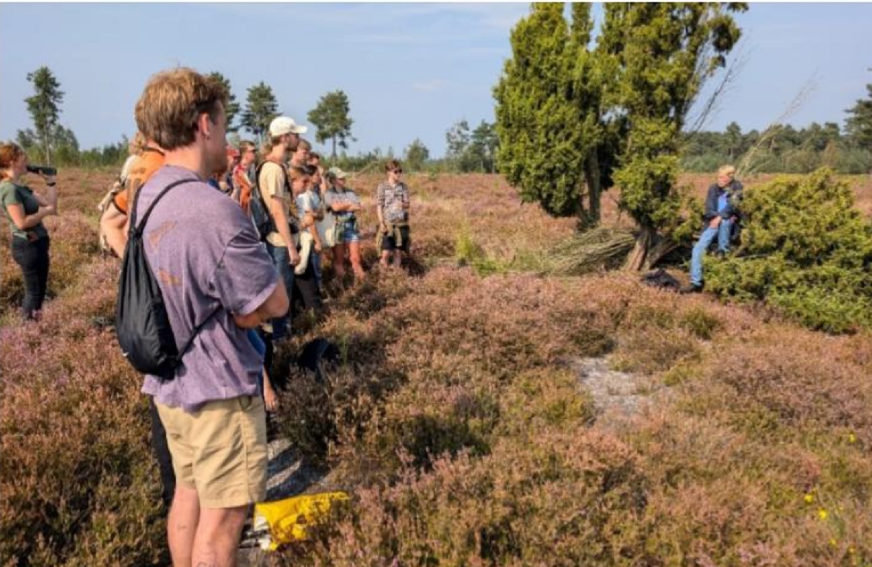 Een groep mensen luistert naar een spreker in een heidelandschap met bomen en struiken.