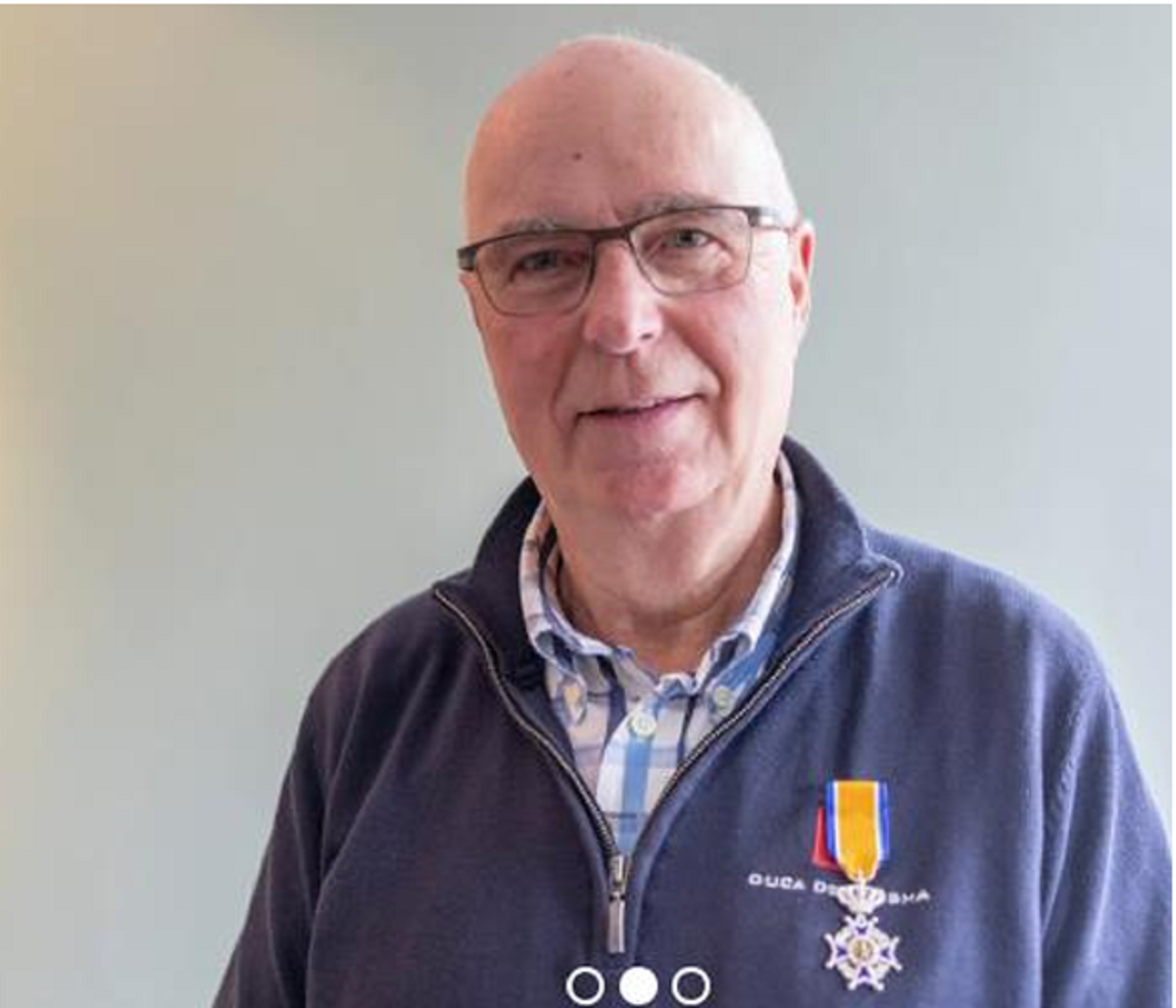 Een man met bril draagt een onderscheiding op zijn trui, tegen een neutrale achtergrond.