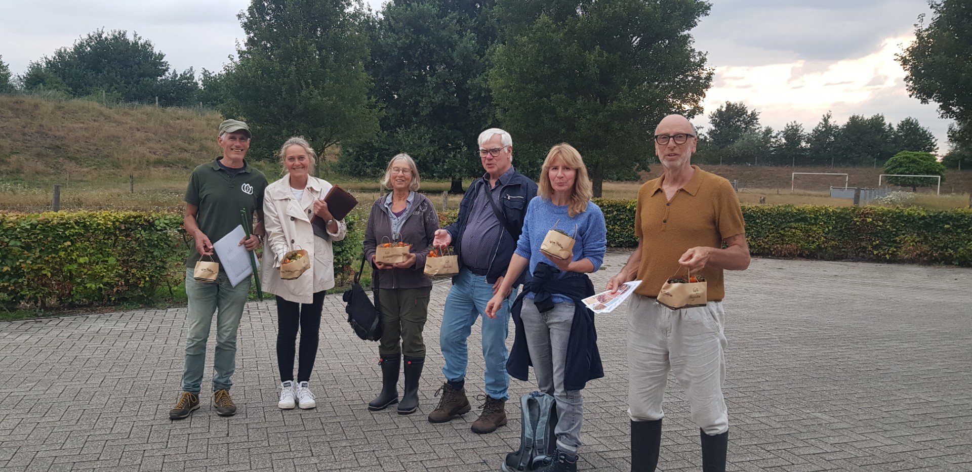 Groep mensen met gevulde mandjes in park, bomen op achtergrond, vroeg in de avond.