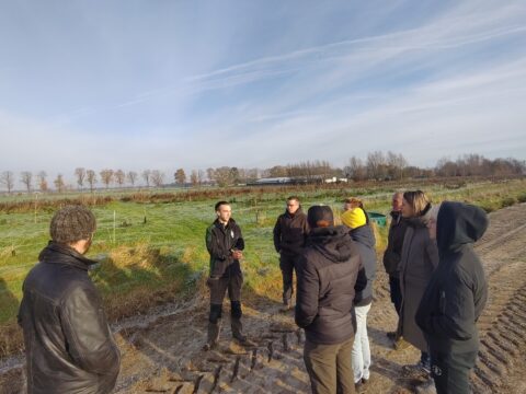 Een groep mensen luistert naar een spreker op een open veld onder een bewolkte hemel.