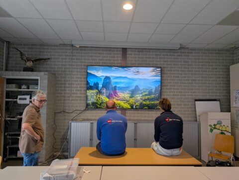 Drie personen kijken naar een grote tv met een natuurlandschap in een kamer met grijze muren.