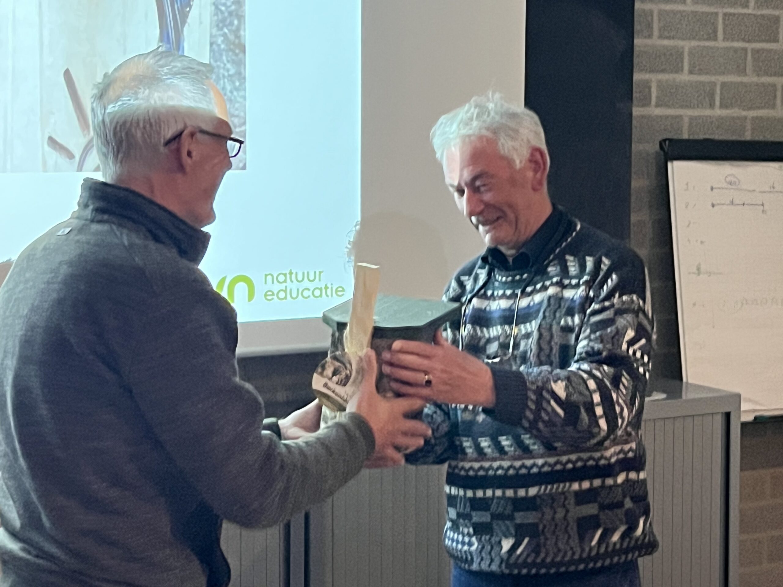 Twee mannen wisselen een houten object uit bij een presentatie over natuureducatie.