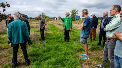 Een groep mannen staat in een grasveld, één wijst naar de verte terwijl anderen luisteren.
