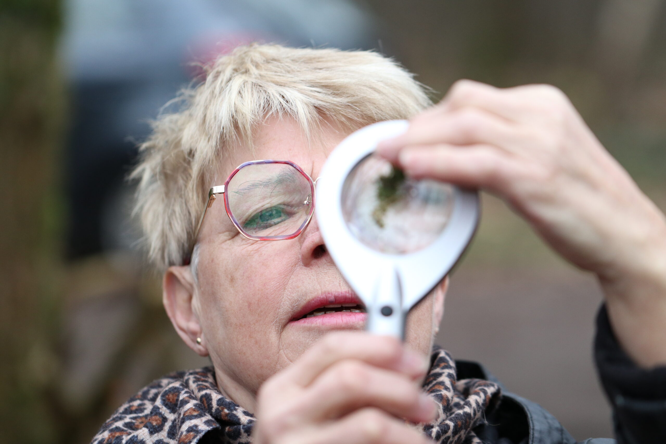Een vrouw met bril bekijkt iets door een loep in een buitenomgeving.