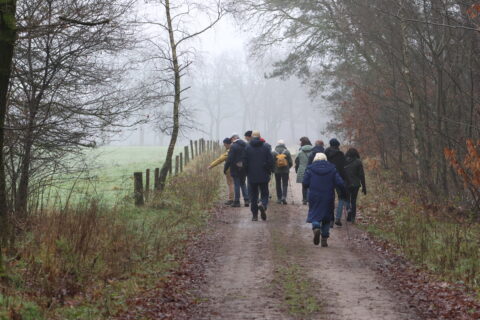 Groep mensen wandelt in mistig bos op modderig pad, omringd door kale bomen.
