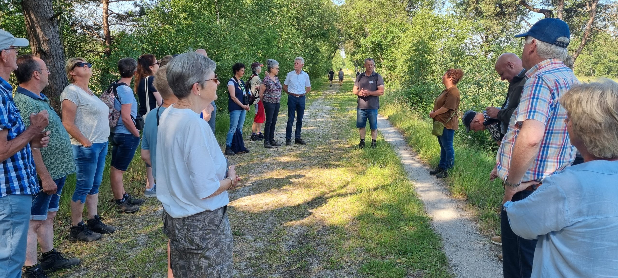 Een groep mensen luistert naar een spreker op een bosrijk pad op een zonnige dag.