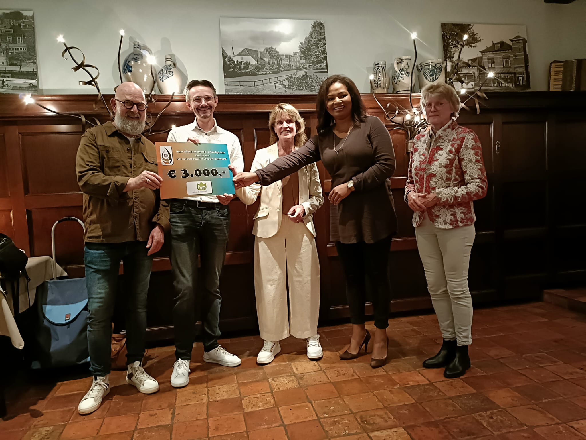 Vijf mensen met een grote cheque van €3000 in een kamer met houten panelen en schilderijen.