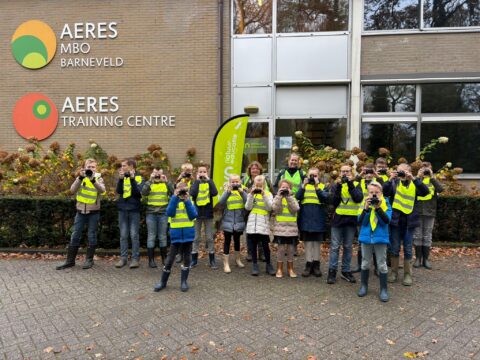 Groep kinderen met verrekijkers en veiligheidsvesten voor het Aeres MBO Barneveld gebouw.