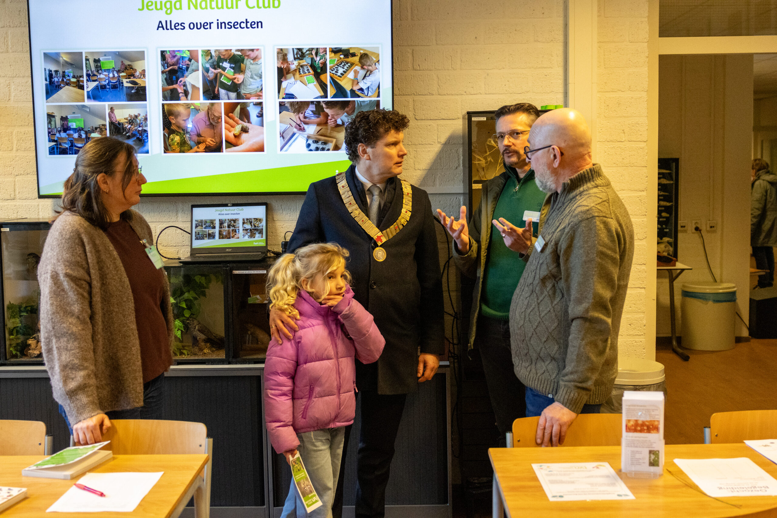 Groep mensen in discussie bij Jeugd Natuur Club presentatie over insecten, met scherm op de achtergrond.