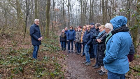 Groep mensen luistert naar een man die een lezing geeft in een bosrijke omgeving.