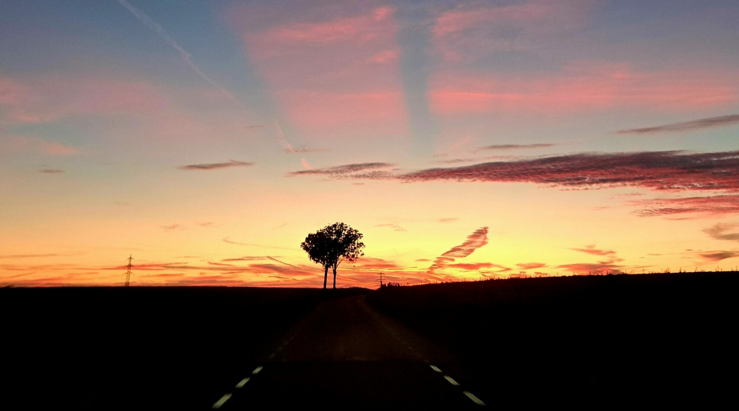 Zonsondergang met silhouet van boom langs een rustige weg, omgeven door kleurrijke wolken.