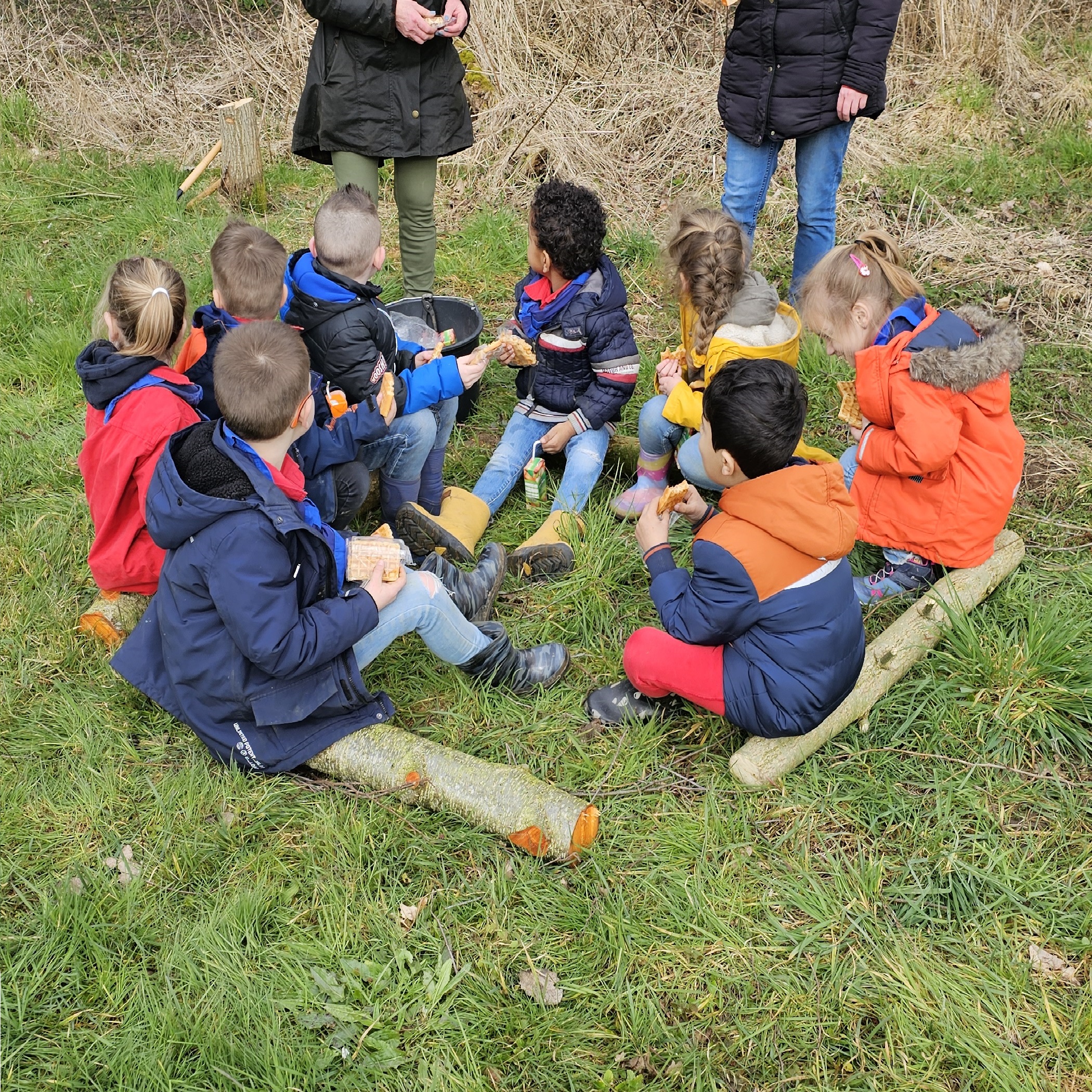 Kinderen zitten in een kring buiten op gras, luisteren en eten een snack.