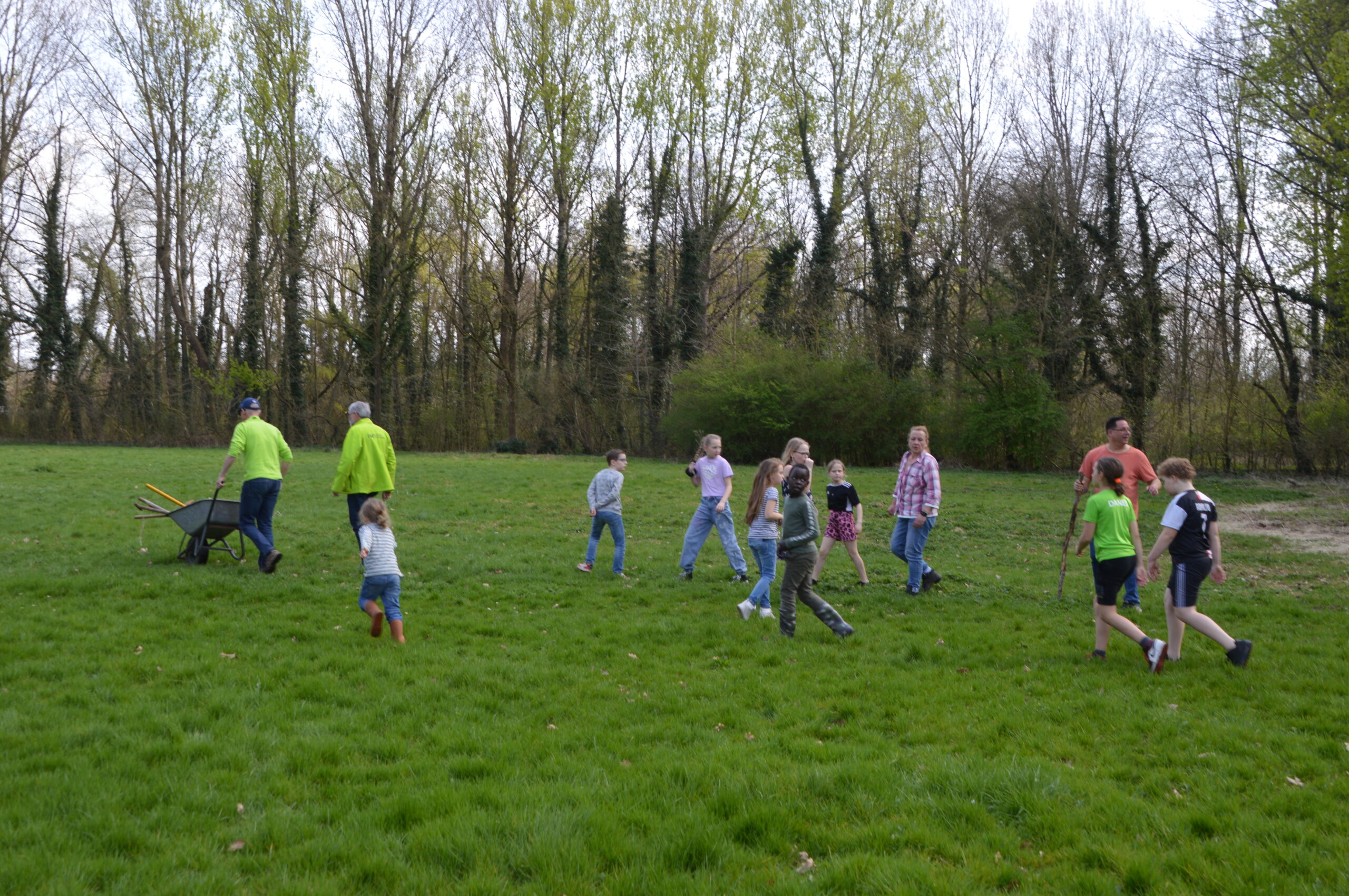 Groep volwassenen en kinderen loopt over grasveld bij bosrand, man duwt kruiwagen met gereedschap.