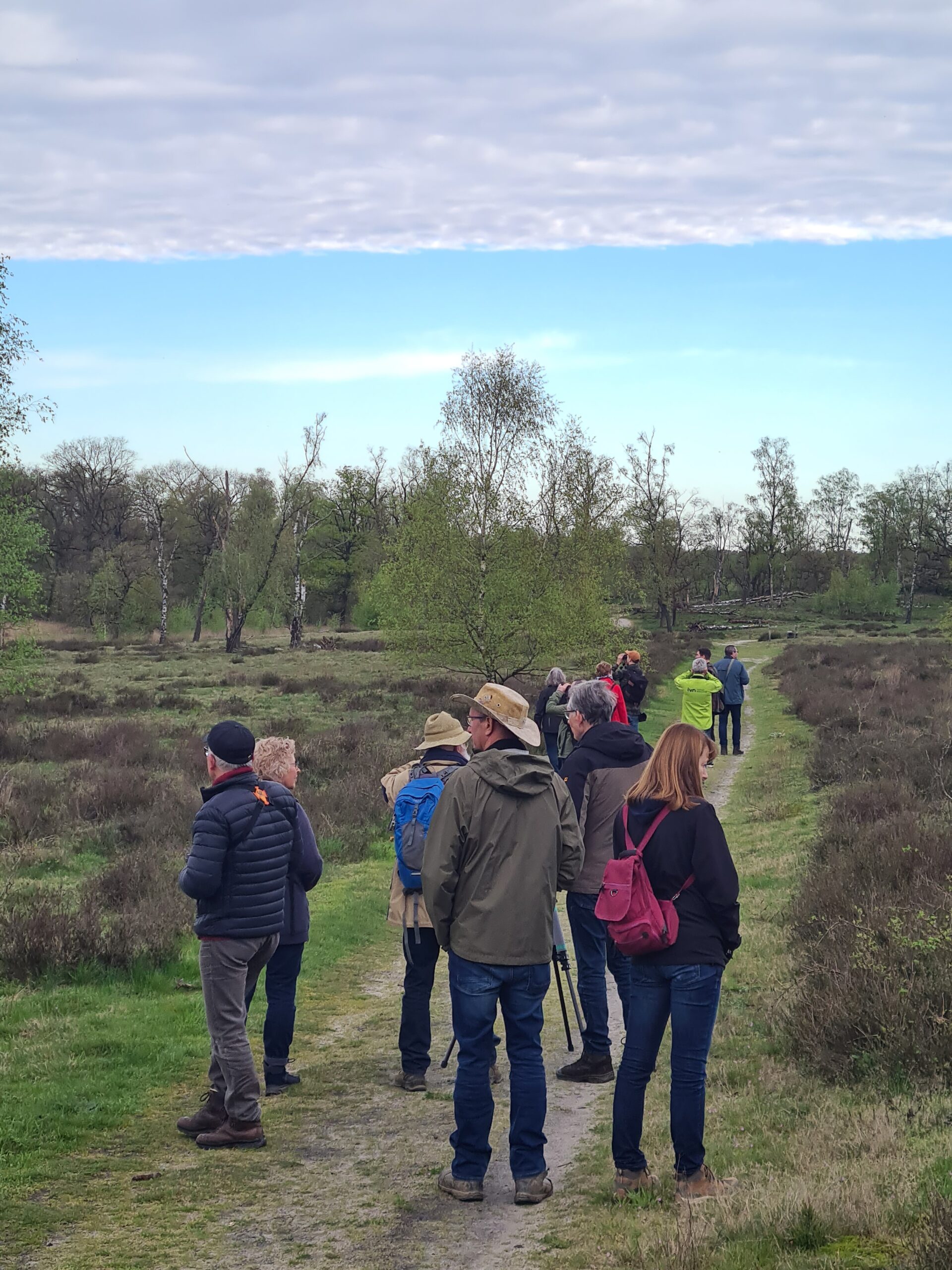 Groep mensen wandelt op een paadje in natuuromgeving onder een bewolkte lucht.