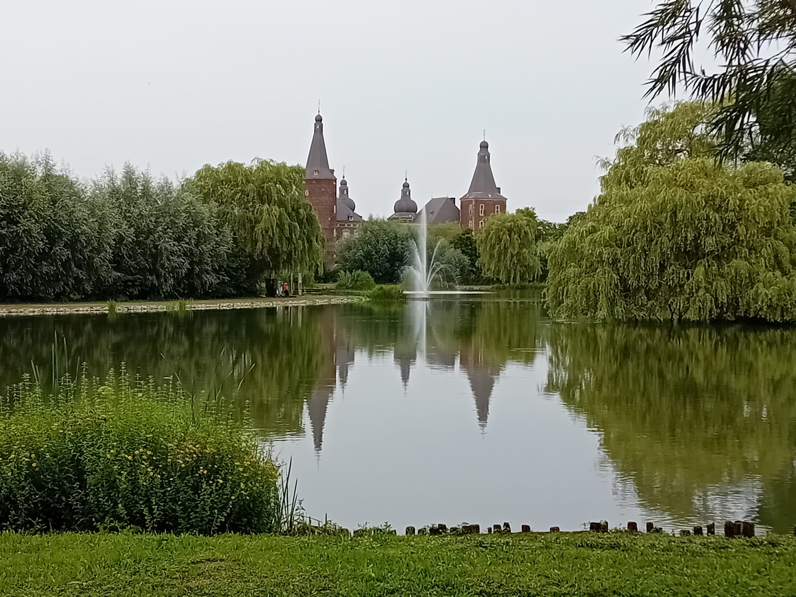 Kasteel met torens weerspiegeld in vijver, omringd door bomen en een fontein in het midden.