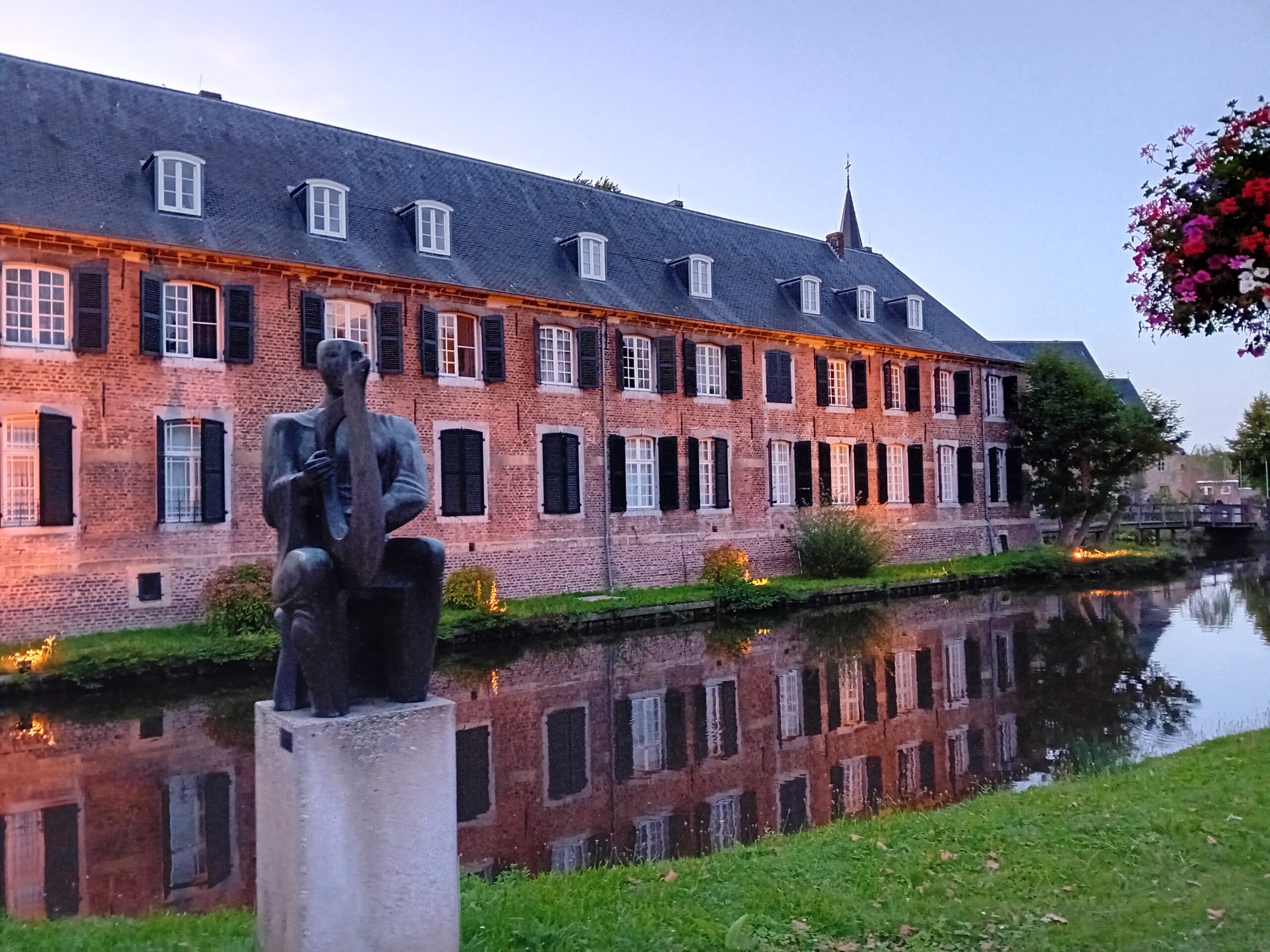 Bakstenen gebouw met zwart-witte luiken weerspiegelt in water; beeldhouwwerk op de voorgrond, bloembak rechts.