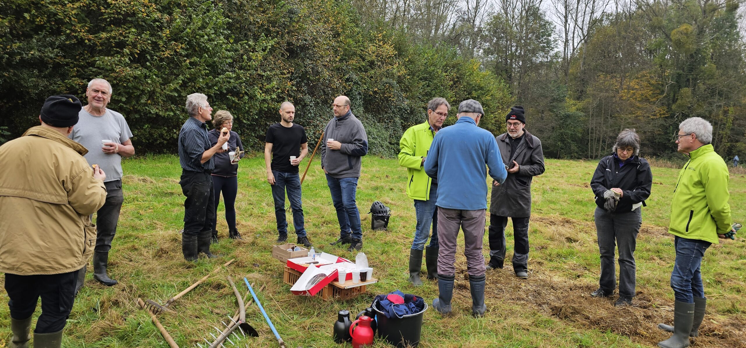 Groep mensen in een veld, genietend van een pauze met drankjes en snacks; herfstbomen op achtergrond.