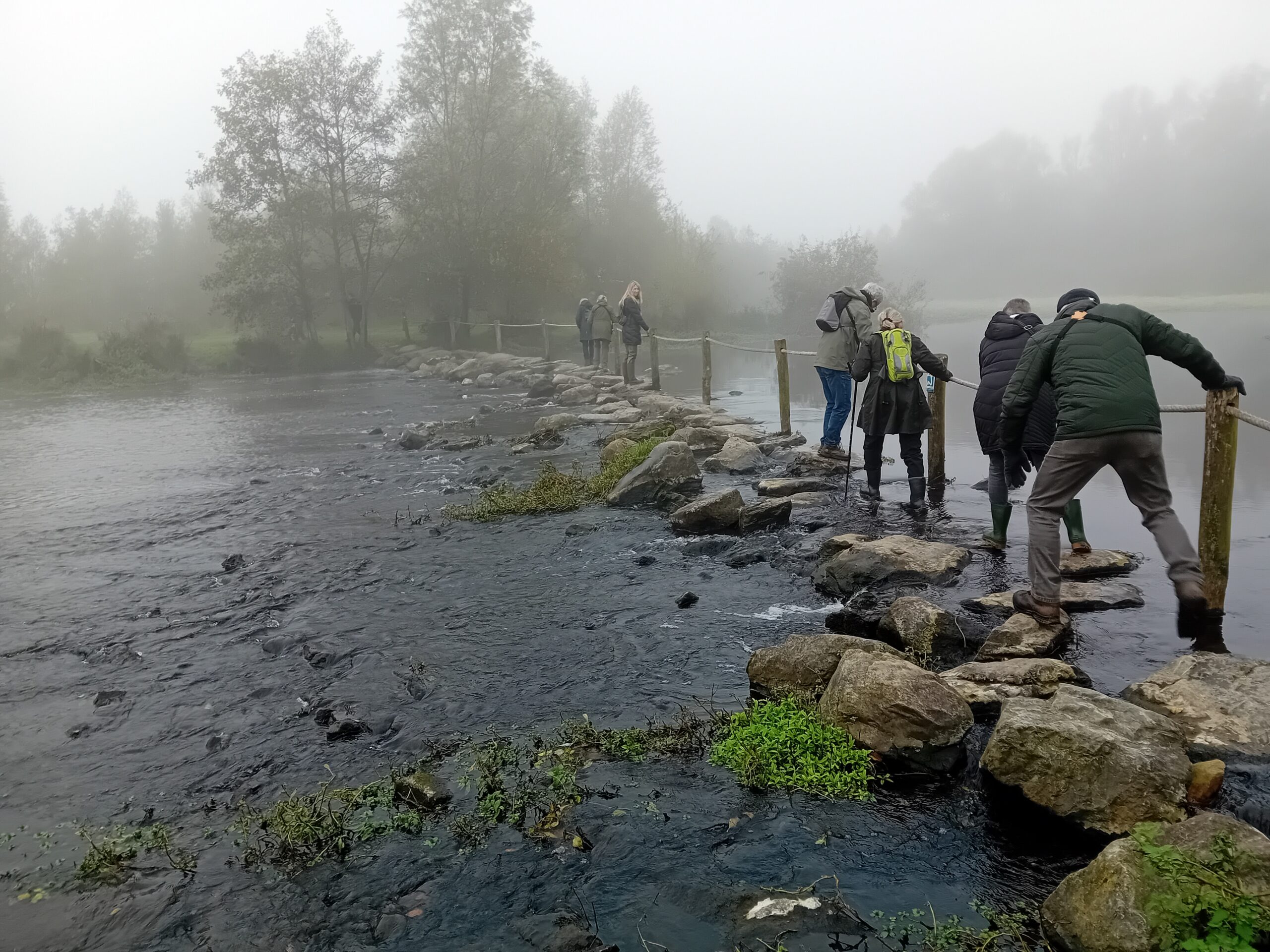 Wandelaars steken een mistige rivier over via rotsblokken en een touwleuning.