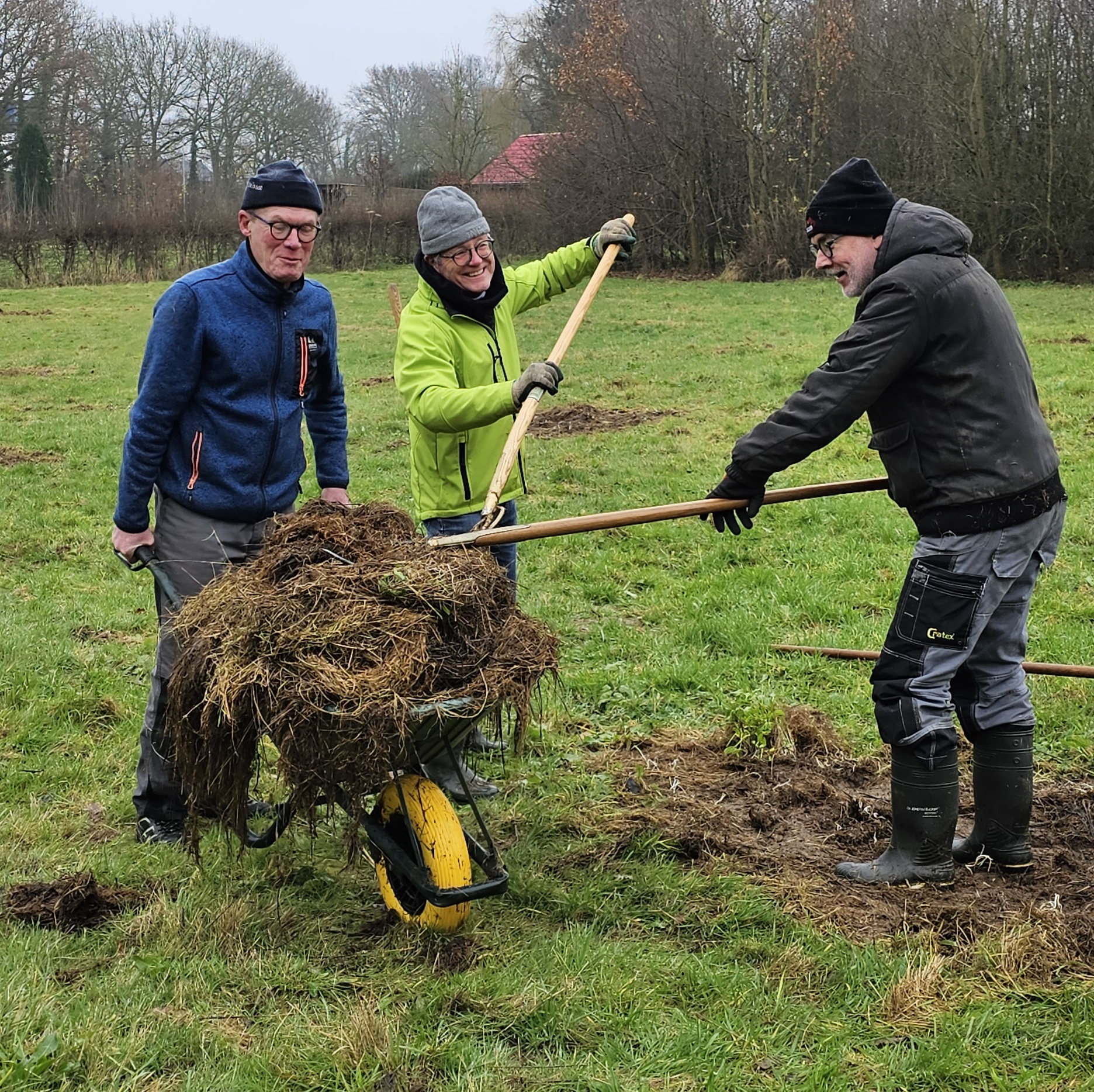 Drie mensen werken samen in een grasveld met gereedschap en een kruiwagen vol tuinafval.