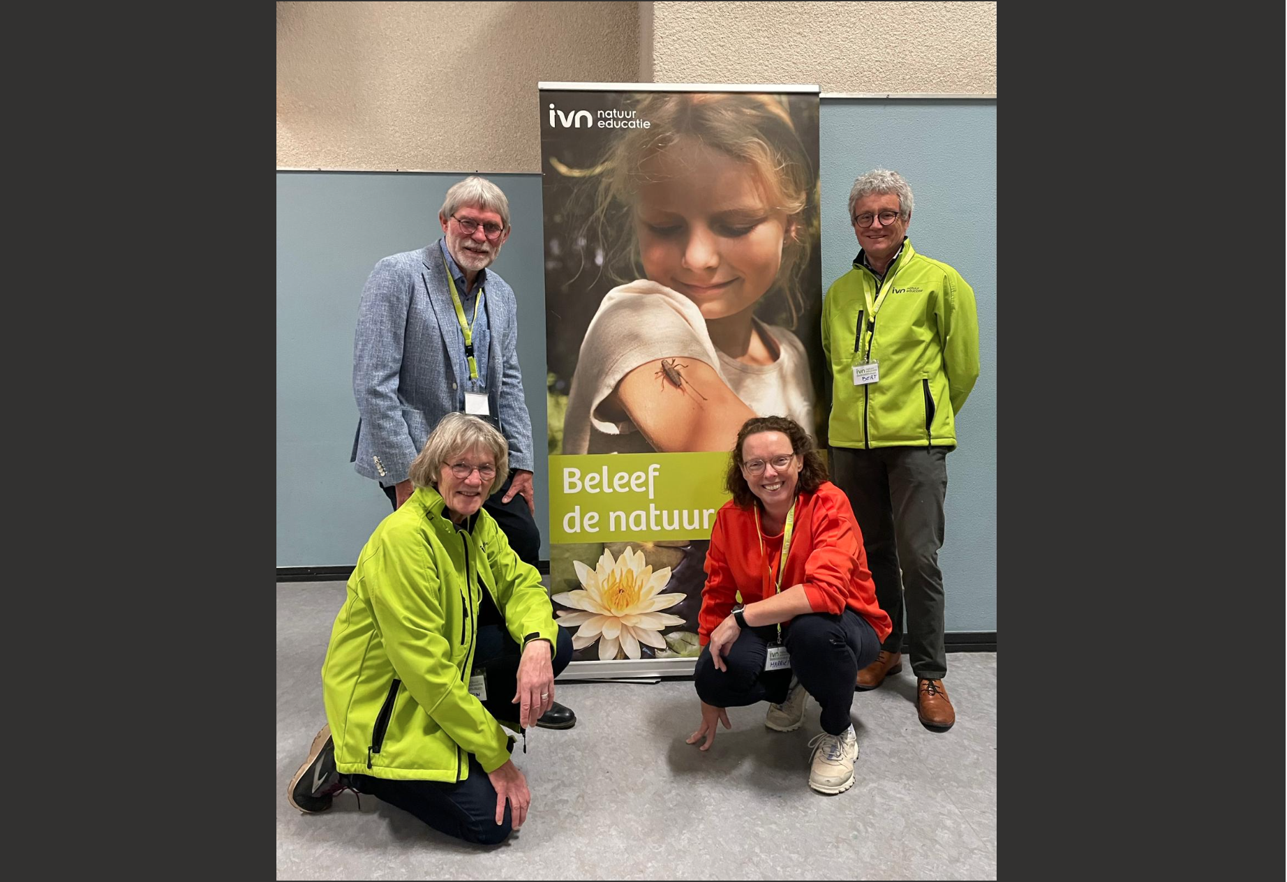 Vier mensen poseren voor een banner met de tekst "Beleef de natuur" in een binnenruimte.
