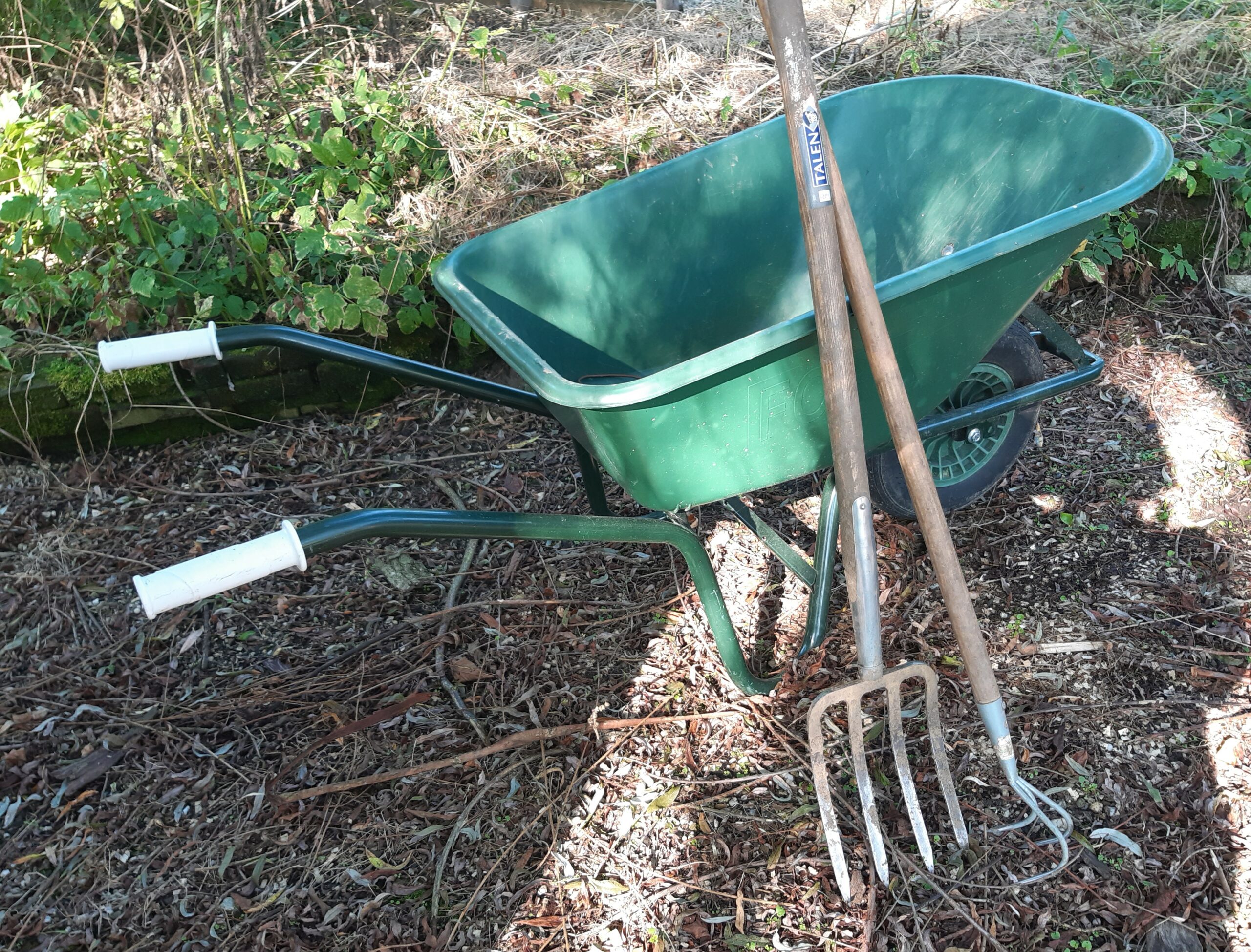 Groene kruiwagen en gereedschap, zoals een hooivork, staan op bruine aarde naast groene planten.