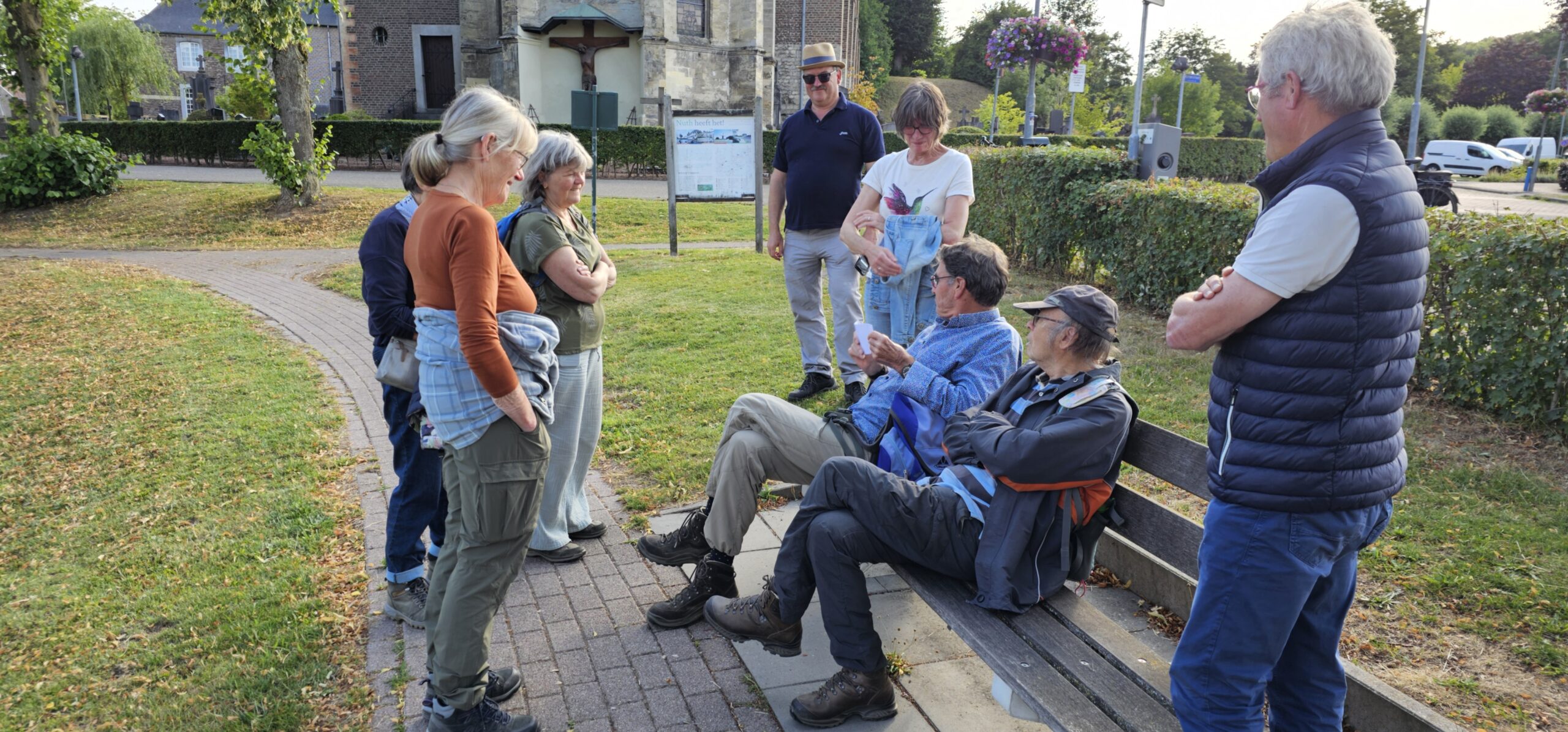 Groep mensen in gesprek in een park, sommigen zitten op een bank, anderen staan.