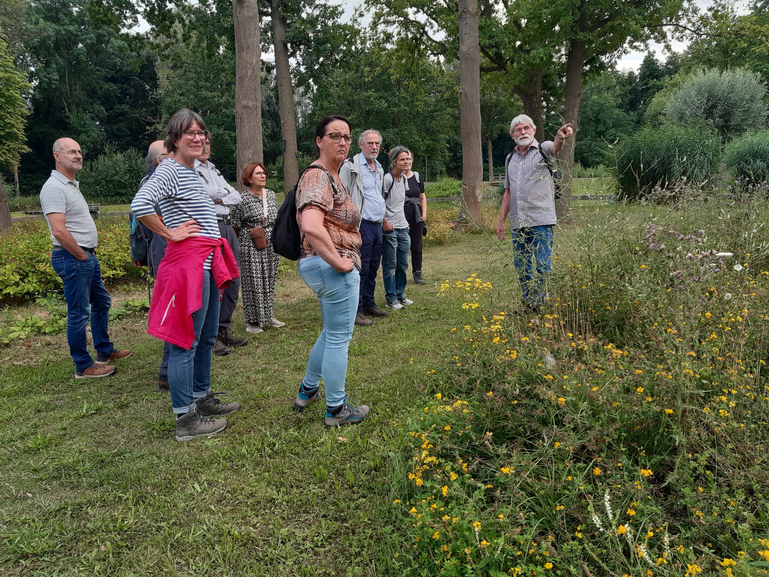 Groep mensen luistert naar een gids in een park met bloemen en bomen op de achtergrond.