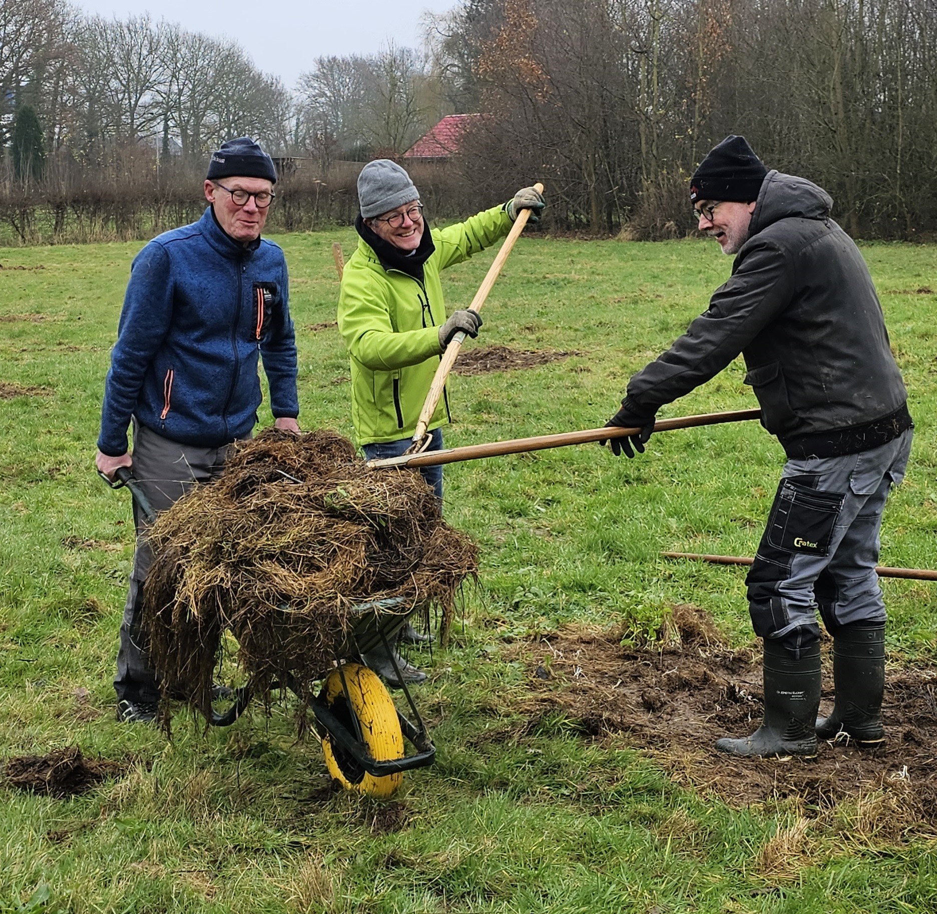 Drie mensen werken samen in een veld, duwen een kruiwagen vol tuinresten.