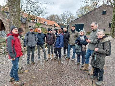 Groep mensen in winterkleding op een boerenerf met bakstenen gebouwen en kale bomen.