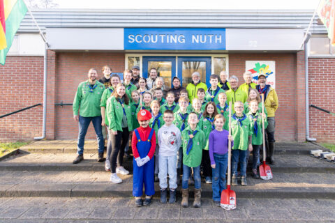 Groep mensen in scoutinguniform voor een gebouw met het bord "Scouting Nuth". Sommige kinderen hebben scheppen.
